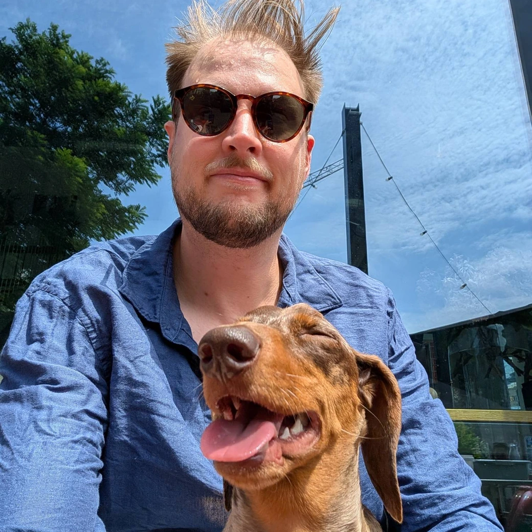 Ali in sunglasses on a sunny terrace, with his dachshund grinning in the foreground.